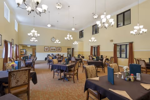 A spacious dining room with multiple tables covered in dark tablecloths, each set with menus, napkins, and condiments. The room features high ceilings with several chandeliers and pendant lights, large windows with brown curtains, and framed artwork on the walls. The carpet is patterned in warm tones, and the overall ambiance is warm and inviting.
