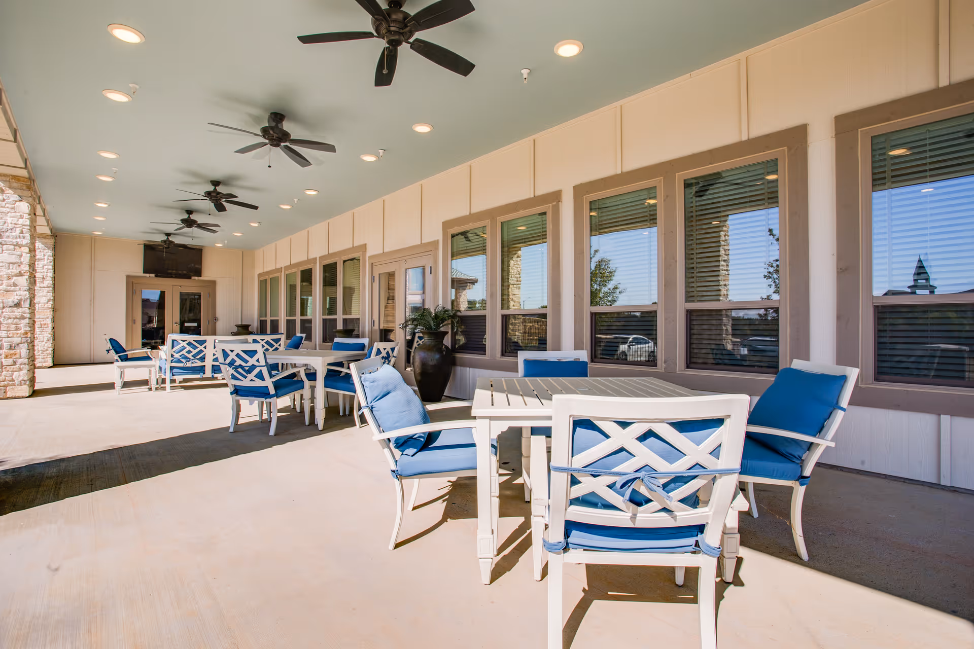 Outdoor covered patio area with several white tables and chairs featuring blue cushions. The ceiling has multiple ceiling fans and recessed lighting. Large windows and a door line the wall on one side, with stone pillars on the other side.
