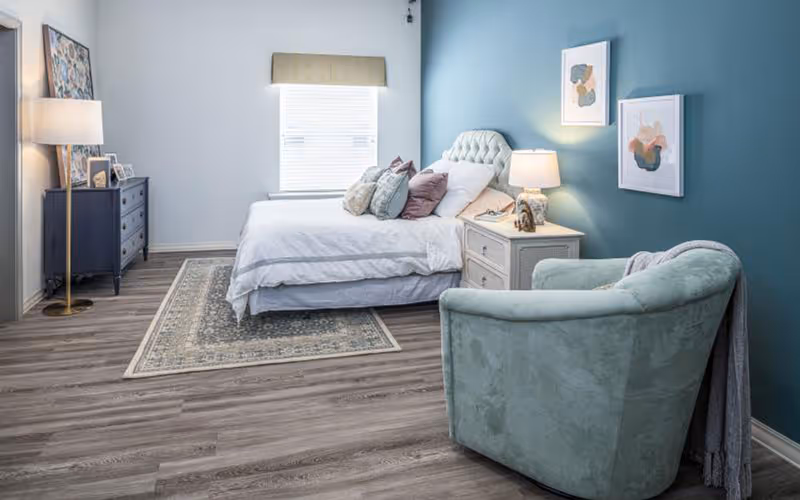 Bedroom with a made bed and tufted headboard, nightstand with lamp, dresser, and a green upholstered chair against a teal accent wall.