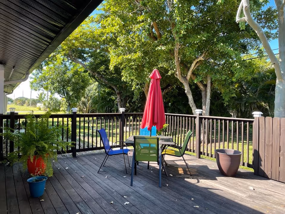 Outdoor wooden deck area with a black railing, featuring a table with a closed red umbrella and four colorful chairs (blue, green, and yellow). There are potted plants on the deck and lush green trees and grass in the background under a clear blue sky.