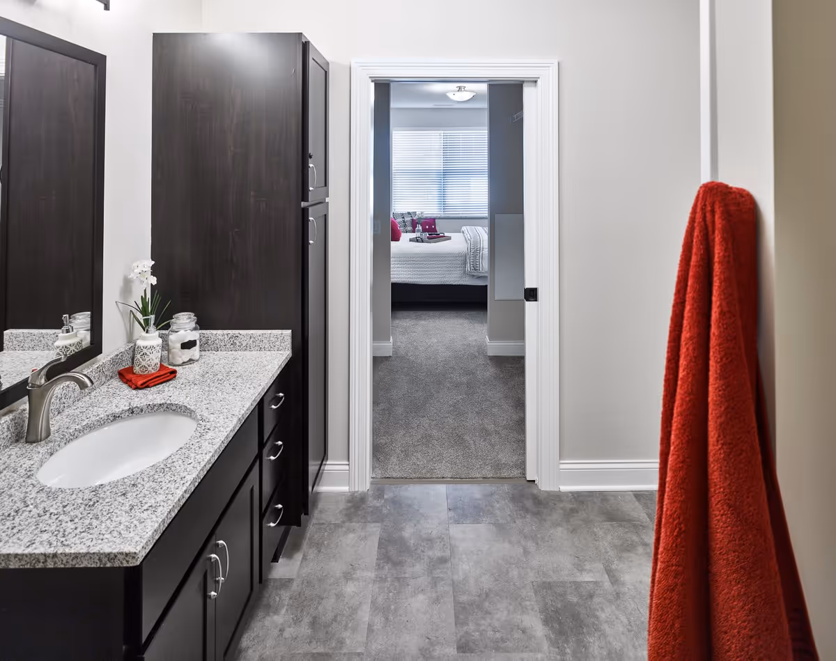 View of a bathroom with a granite countertop, dark wood cabinets, a sink, and a mirror on the left side. A red towel hangs on a hook on the right wall. Through an open doorway, a bedroom with a bed, white bedding, and a window with blinds is visible.