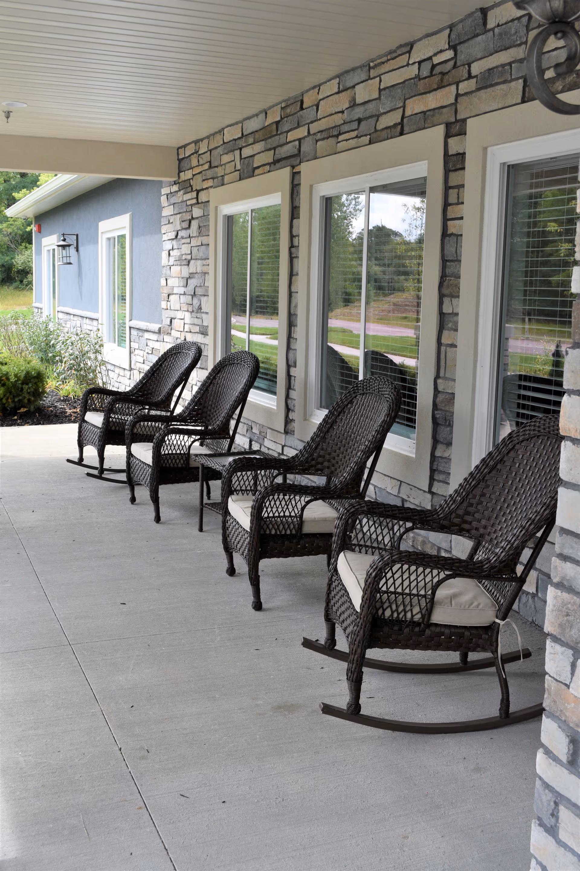 A covered outdoor porch area with four dark wicker rocking chairs with light cushions lined up against a stone and stucco wall with windows reflecting greenery outside.