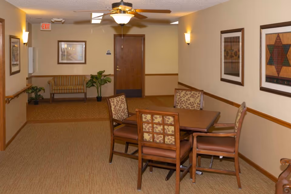 Indoor common area of a senior living facility with a table and four chairs, framed artwork on the walls, and a bench in the background.