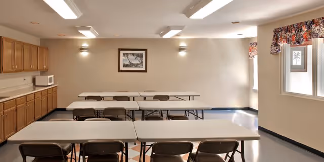 Interior view of a room with several rectangular tables and folding chairs arranged in rows. The room has beige walls, wooden cabinets along one side with a microwave on the countertop, and two windows with floral valances. A framed picture hangs on the wall, and ceiling lights illuminate the space.