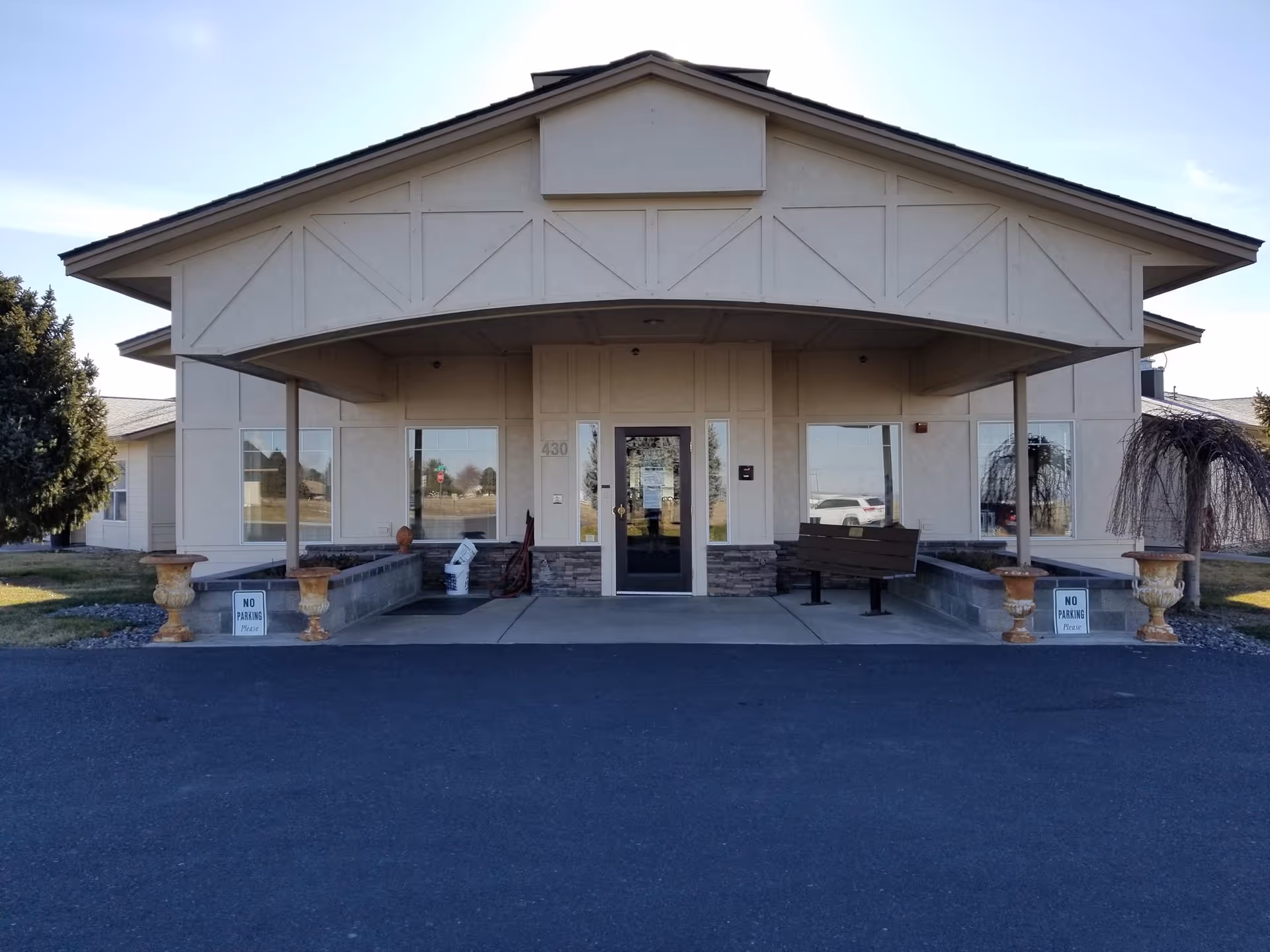 Front exterior view of Coventry House Assisted Living building with a covered entrance, two large windows on either side of the door, a bench on the right side, and decorative planters near the entrance.