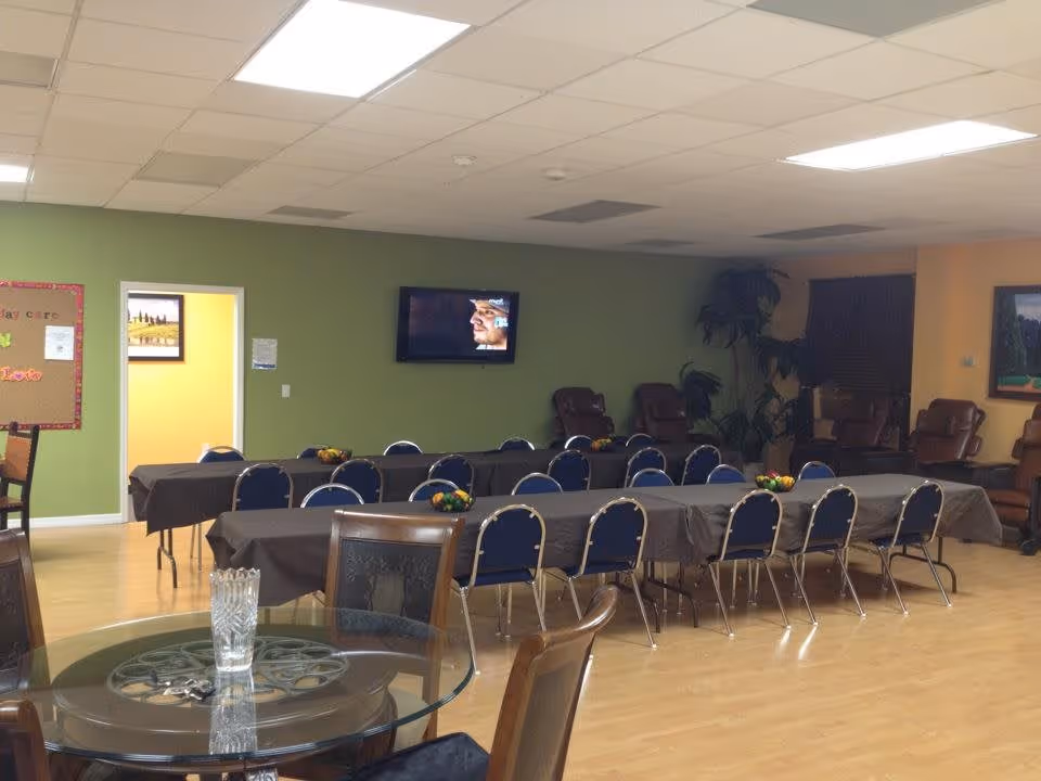 A spacious room with several tables covered in dark tablecloths, surrounded by blue chairs. A glass round table with wooden chairs is in the foreground. The walls are painted green and yellow, with a flat-screen TV mounted on the green wall showing a person's face. There are some plants and brown recliner chairs along the back wall, and a bulletin board is visible on the left side.