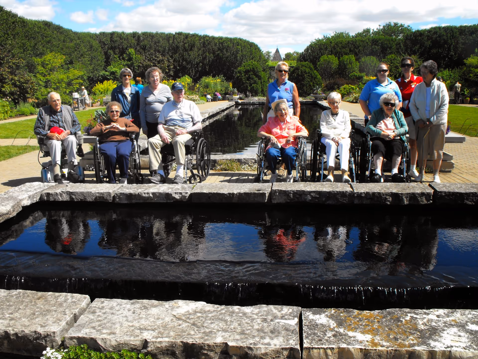 A group of elderly people, some in wheelchairs and some standing, posing together outdoors in front of a long rectangular water feature with greenery and trees in the background under a partly cloudy sky.