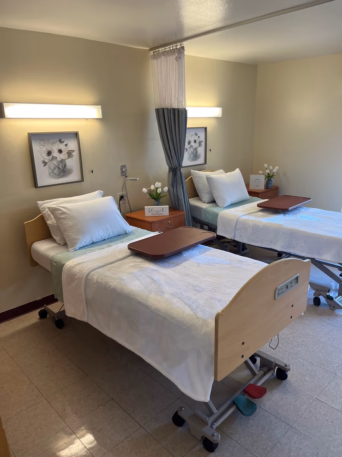 A room with two hospital-style beds each with white pillows and blankets, wooden headboards and footboards, and brown overbed trays. Between the beds is a small wooden nightstand with a vase of white tulips and a small sign. Above each bed is a wall-mounted light and a framed picture of white flowers in a vase. A privacy curtain is partially drawn between the beds.