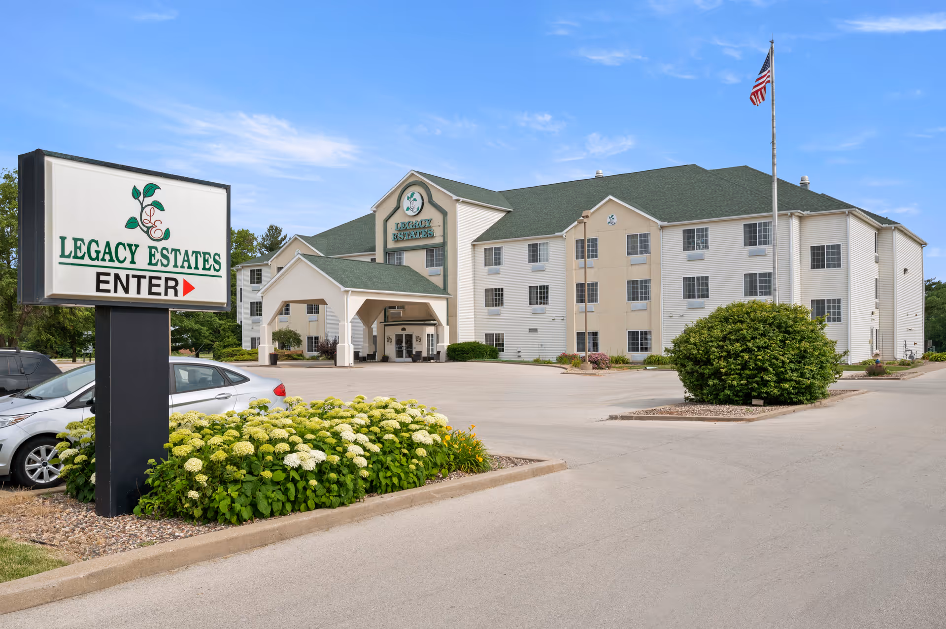 Exterior view of Legacy Estates of Monmouth, a three-story building with a green roof and beige siding. There is a large sign in the foreground that reads 'Legacy Estates ENTER' with an arrow pointing right. The building has a covered entrance, several windows, a flagpole with an American flag, and landscaped areas with bushes and flowers. Several parked cars are visible on the left side.