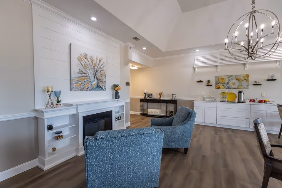 A cozy living room area with two blue upholstered armchairs facing a white fireplace mantel decorated with a peacock painting, candles, and small plants. The room features light wood flooring, a high ceiling with recessed lighting, and a modern chandelier. In the background, there is a white cabinet with a coffee and tea station, decorated with floral artwork and small shelves holding plants and decor items.