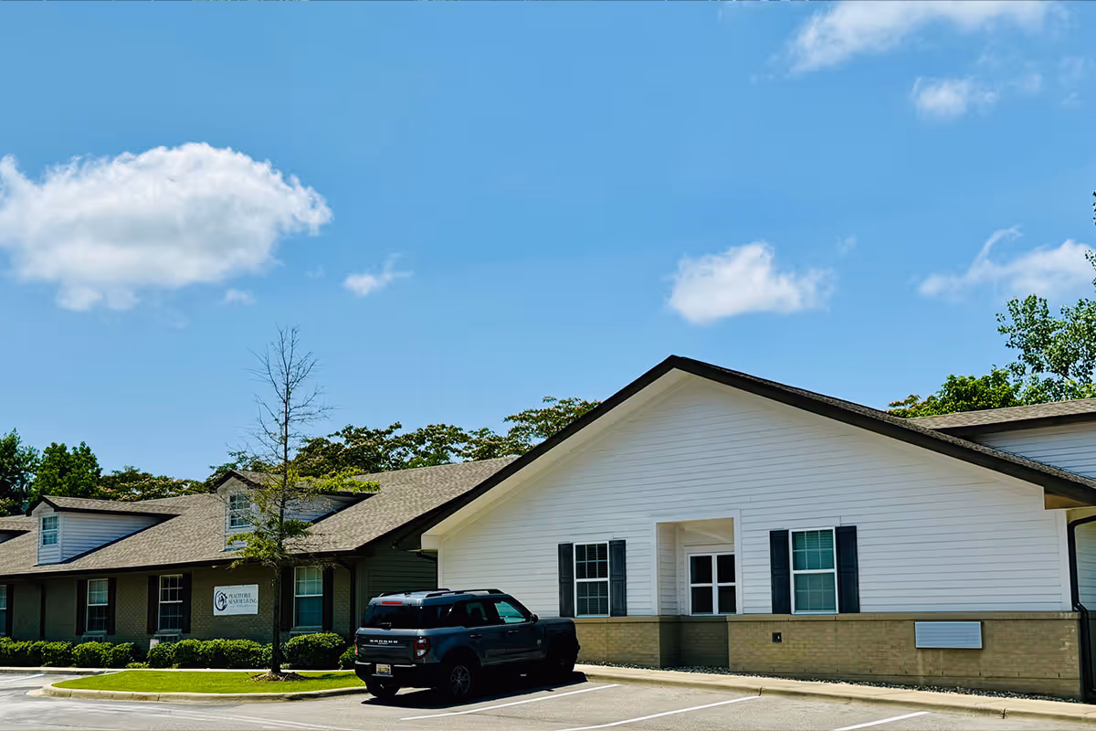Exterior view of Volante Senior Living of Peachtree building under a blue sky with some clouds. The building has white siding with a brick lower section and black shutters on the windows. A black SUV is parked in front of the building in a parking lot.