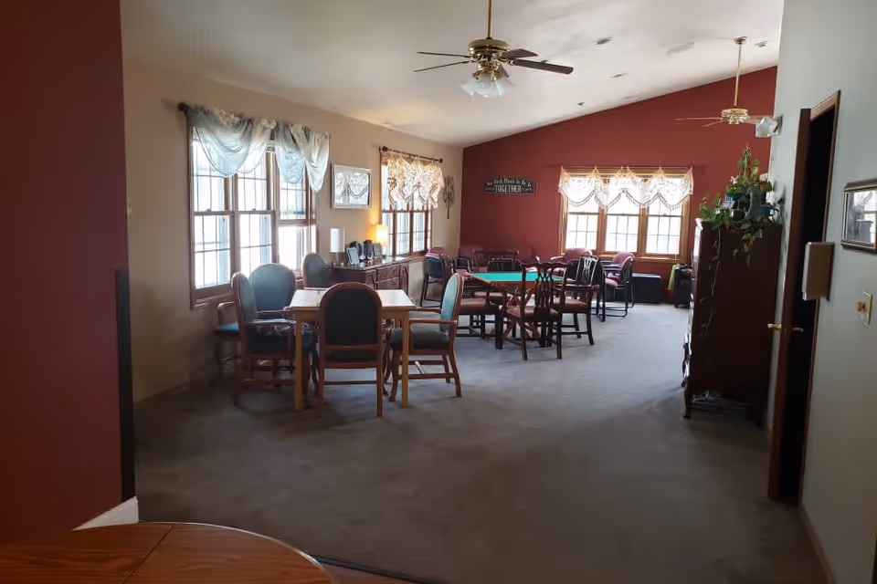 Interior view of a senior living facility common area with multiple tables and chairs arranged for socializing or activities. The room has large windows with sheer curtains allowing natural light, a ceiling fan, and a burgundy accent wall with a decorative sign. There are plants and framed pictures on the walls, creating a cozy and welcoming atmosphere.