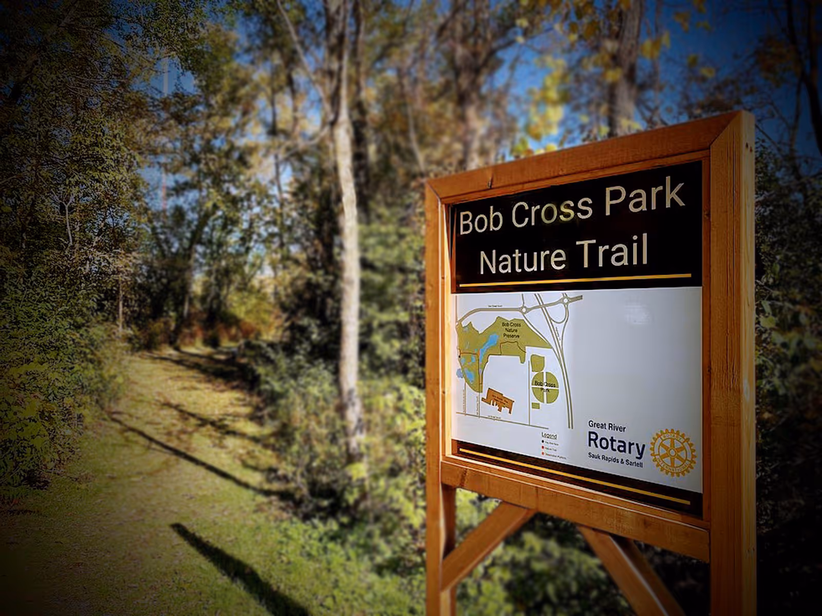 A wooden signboard for Bob Cross Park Nature Trail with a map and the Great River Rotary logo, surrounded by trees and greenery under a clear blue sky.