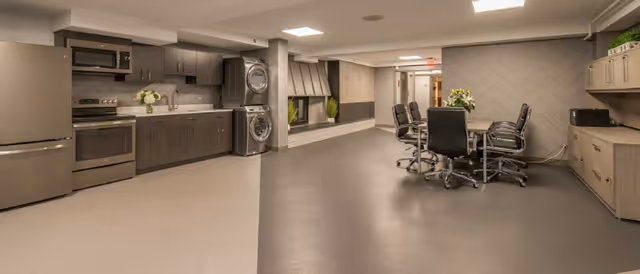 A modern communal room featuring a kitchen area with stainless steel appliances including a refrigerator, oven, microwave, and stacked washer and dryer. Adjacent to the kitchen is a seating area with a table and several office-style chairs. The room has neutral tones with gray cabinetry, light-colored walls, and a patterned accent wall. There are decorative plants and flowers placed on the countertops and table.