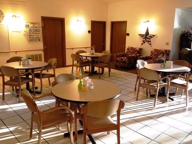 A well-lit dining area with several round tables and chairs arranged neatly. The room has tiled flooring and light-colored walls with three wooden doors on one side. There are two patterned armchairs near the wall with a star decoration above them. Small flower vases and condiments are placed on the tables.