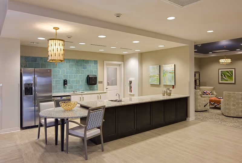 A modern kitchen area with a long granite countertop island, a small round dining table with two chairs, a stainless steel refrigerator, and a turquoise tiled backsplash. The kitchen opens into a living room area with armchairs, a coffee table, and wall art.