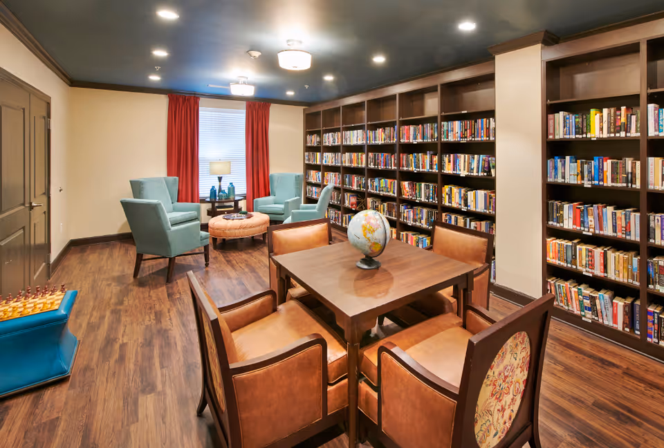 Well-lit community library/reading room with bookshelves lining the walls, a central table with chairs and a globe, and armchairs by a window with red curtains.