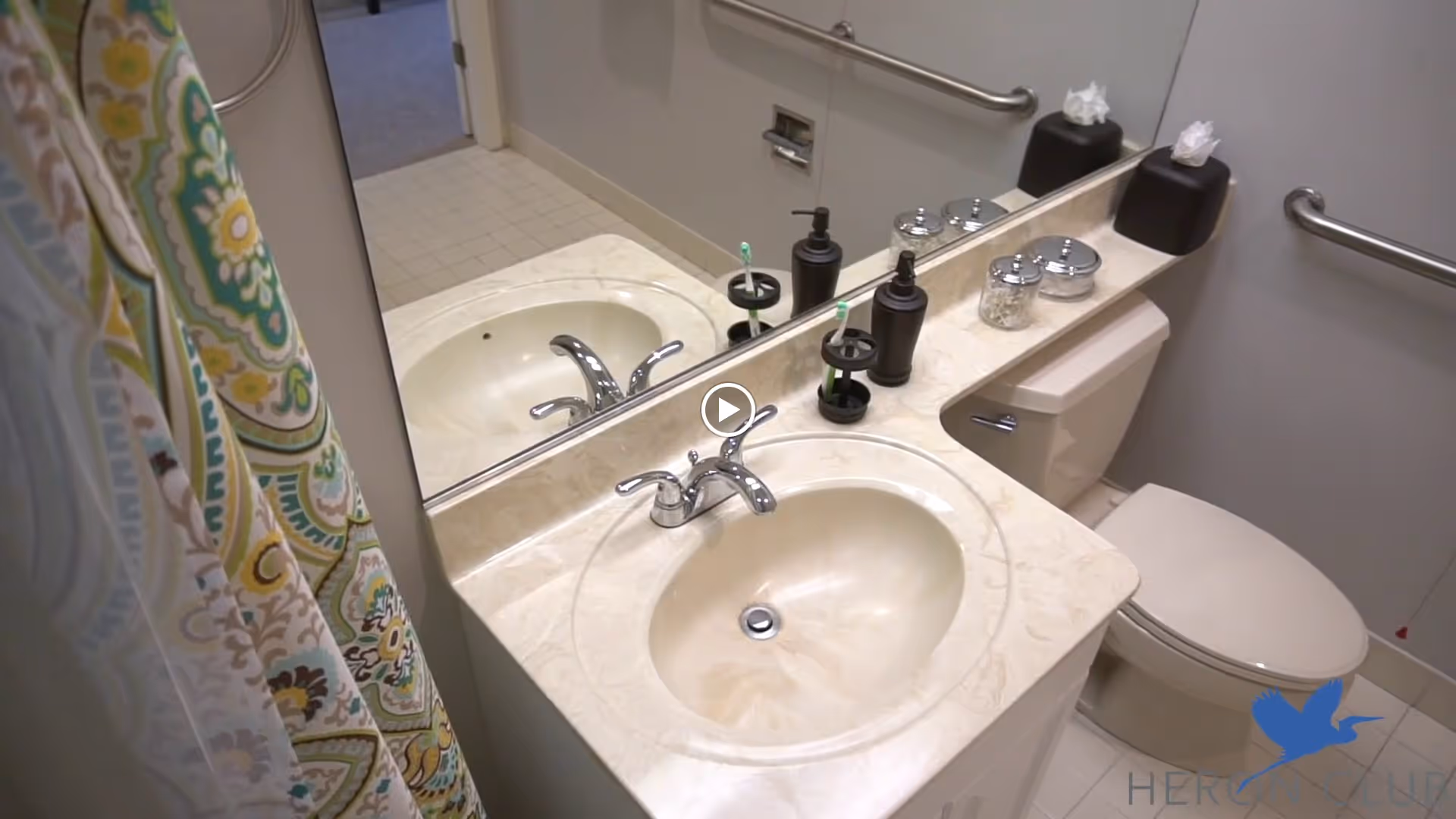 Small accessible bathroom with a sink and countertop, mirror, toilet, grab bars, soap dispensers, and a patterned shower curtain.