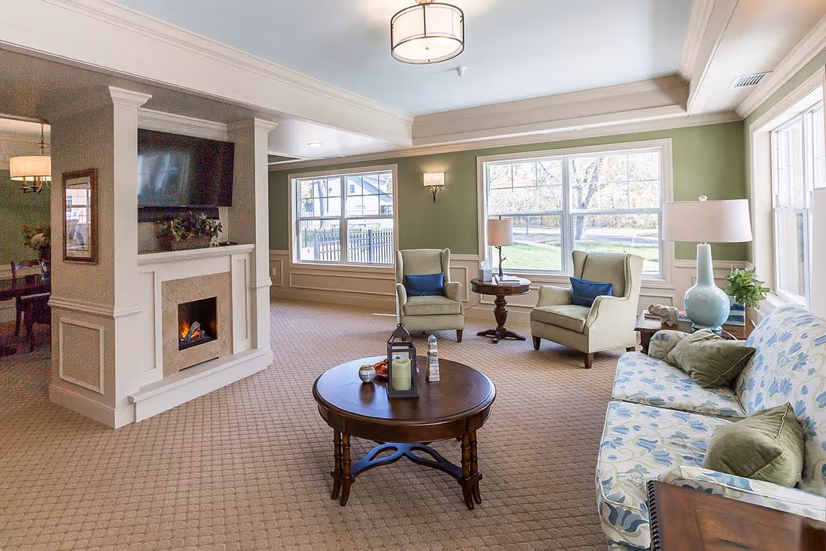 A cozy living room with a beige carpet, two green armchairs with blue cushions, a floral patterned sofa with green pillows, a round wooden coffee table with decorative items, a fireplace with a mounted TV above it, and large windows letting in natural light.