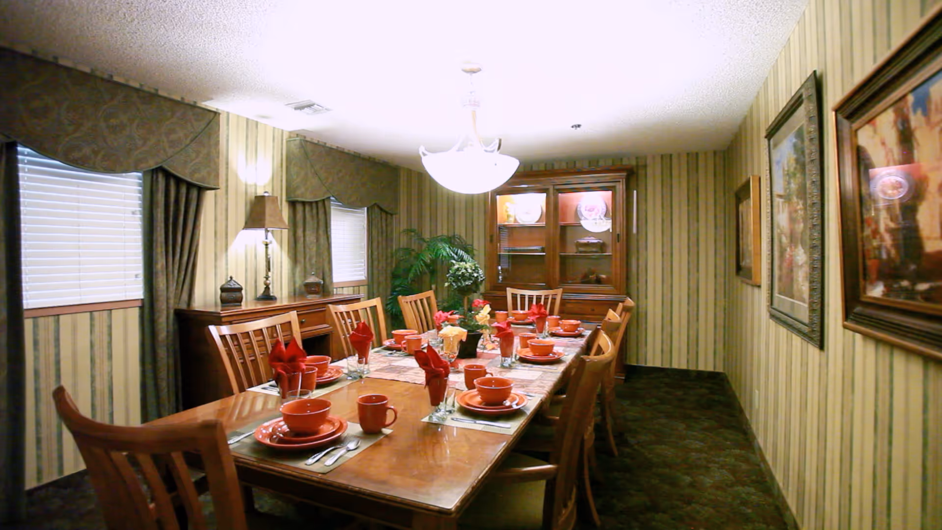 A formal dining room with a long wooden table set for eight people. Each place setting includes red dishes, cups, and folded red napkins. The room has striped wallpaper, two windows with valances and curtains, a wooden sideboard with a lamp and decorative items, a wooden china cabinet with plates on display, and framed artwork on the walls. A ceiling light fixture hangs above the table.