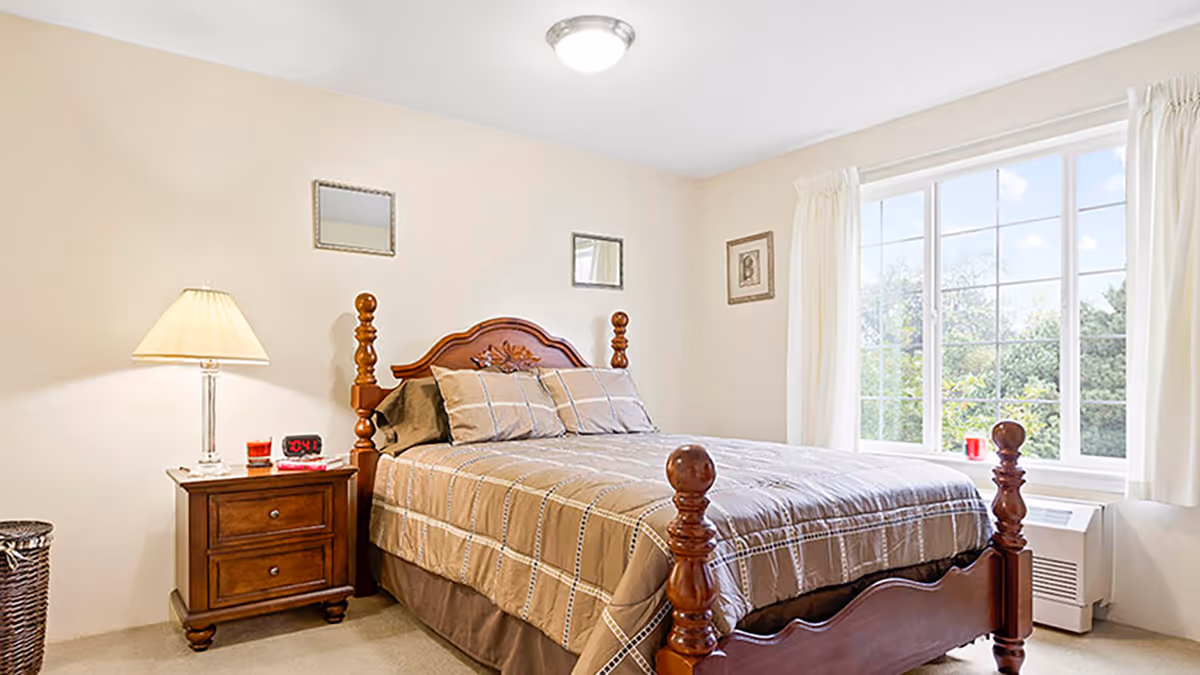 Well-lit bedroom with a wooden four-poster bed, a nightstand with a lamp and alarm clock, and a large window showing greenery outside.