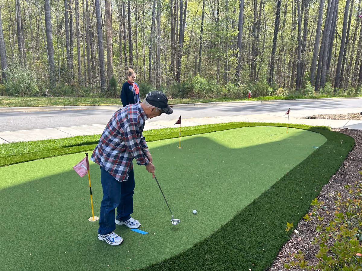 An elderly man wearing a plaid shirt, jeans, and a black cap is putting a golf ball on a small artificial putting green outdoors. A woman stands nearby watching him. The background shows a wooded area with tall trees and a road.