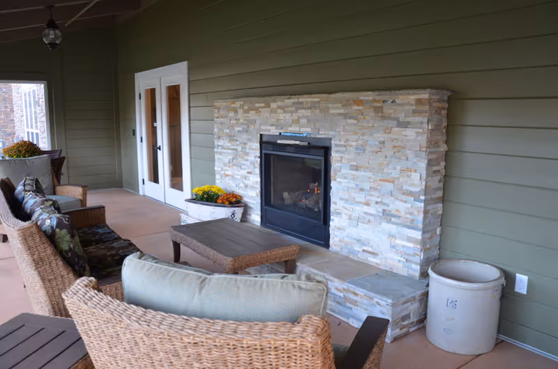 Covered outdoor patio area with wicker chairs and a coffee table facing a stone fireplace built into a green paneled wall. There are potted flowers near the fireplace and double glass doors leading inside.