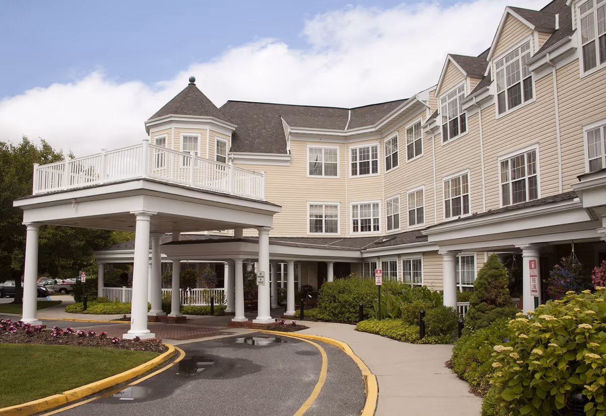 Exterior view of a multi-story senior living facility building with beige siding and white trim. The entrance features a covered drop-off area supported by white columns, surrounded by landscaped greenery and flowers. The sky is partly cloudy.