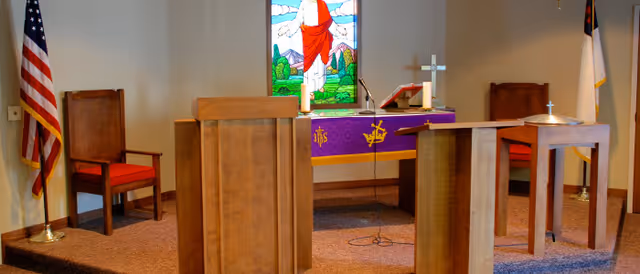 Small interior chapel with wooden pulpit and altar, a stained-glass window depicting Jesus, chairs, and flags.