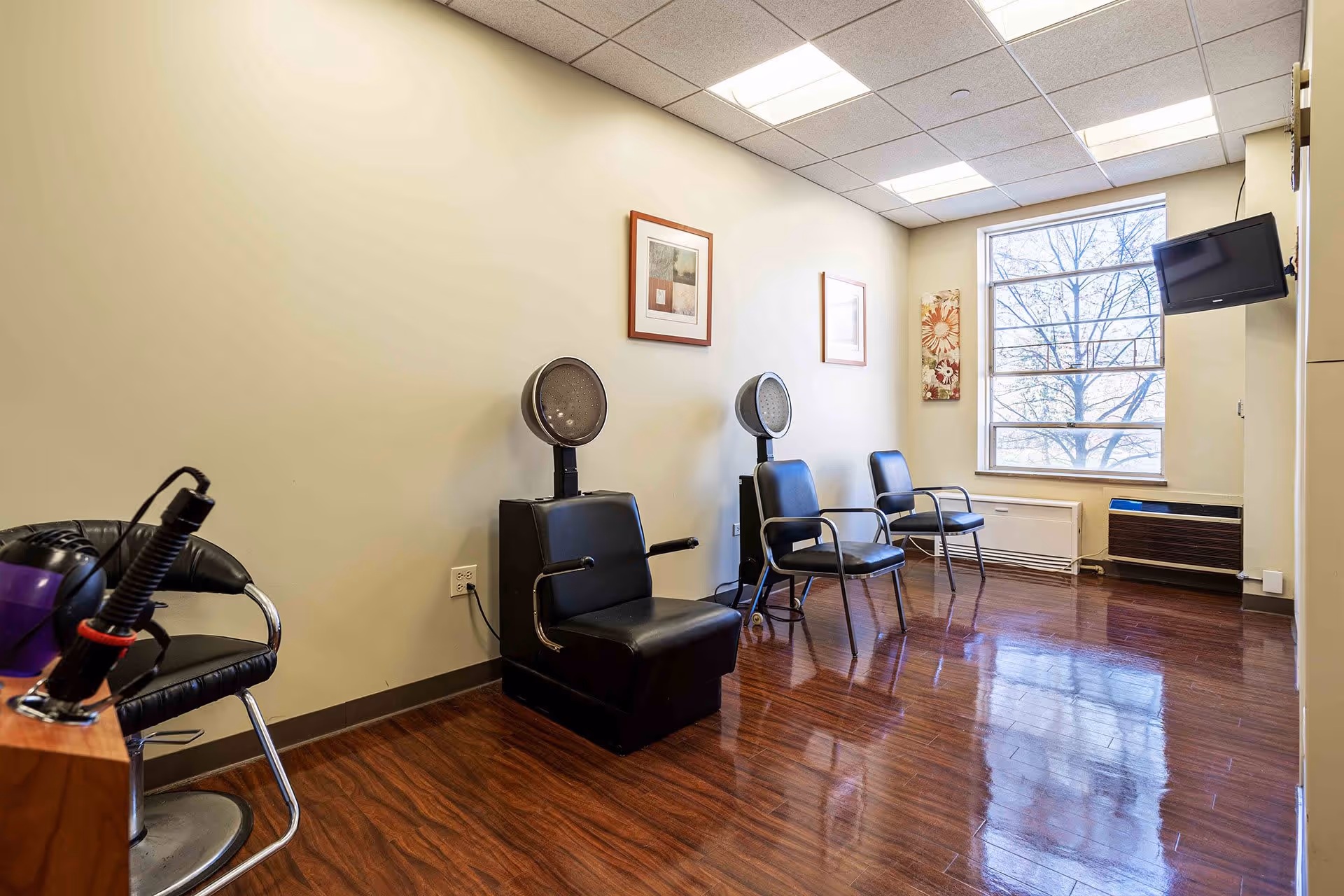 Interior view of a hair salon area in a senior living facility with three black salon chairs, two hair dryers, a large window letting in natural light, framed artwork on the walls, and a mounted TV in the corner.