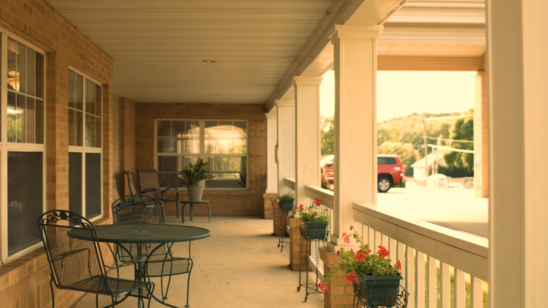 Covered brick porch with a metal patio table and chairs, potted plants along the railing and a red SUV parked in the background.