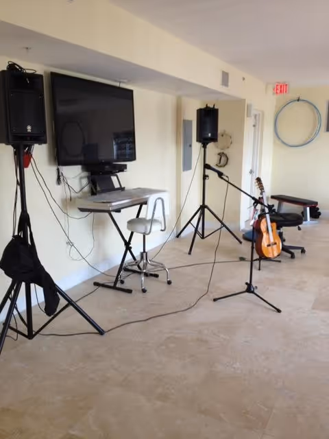 A room with musical equipment including a keyboard on a stand with a chair, two large speakers on tripods, a microphone on a stand, and an acoustic guitar resting on a chair. The room has beige tiled flooring and light-colored walls with an exit sign above a door in the background.
