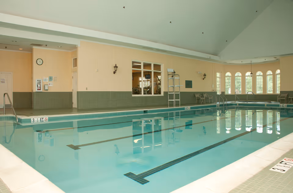 Indoor swimming pool with clear blue water, surrounded by beige and green tiled walls. There are several metal ladders for pool access, chairs along the far wall, and large windows letting in natural light. A clock and some safety signs are visible on the walls.