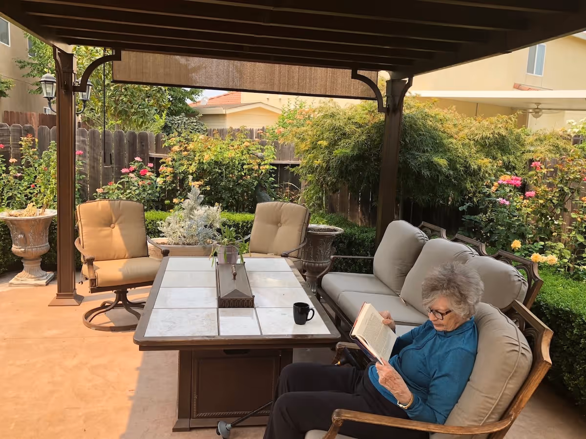 An elderly woman with gray hair and glasses is sitting on a cushioned outdoor sofa under a pergola, reading a book. The patio area features a tiled table with a small plant and a coffee mug on it, surrounded by cushioned chairs. The background shows a wooden fence with various green bushes and blooming flowers.