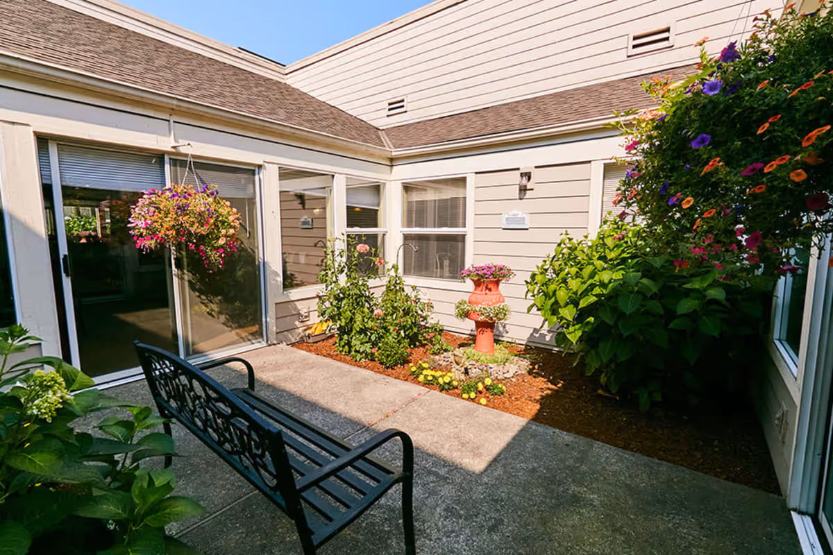 A small outdoor courtyard area with a black metal bench, hanging flower baskets, and various plants and flowers along the edges. The courtyard is surrounded by beige siding walls with windows and a sliding glass door leading inside.