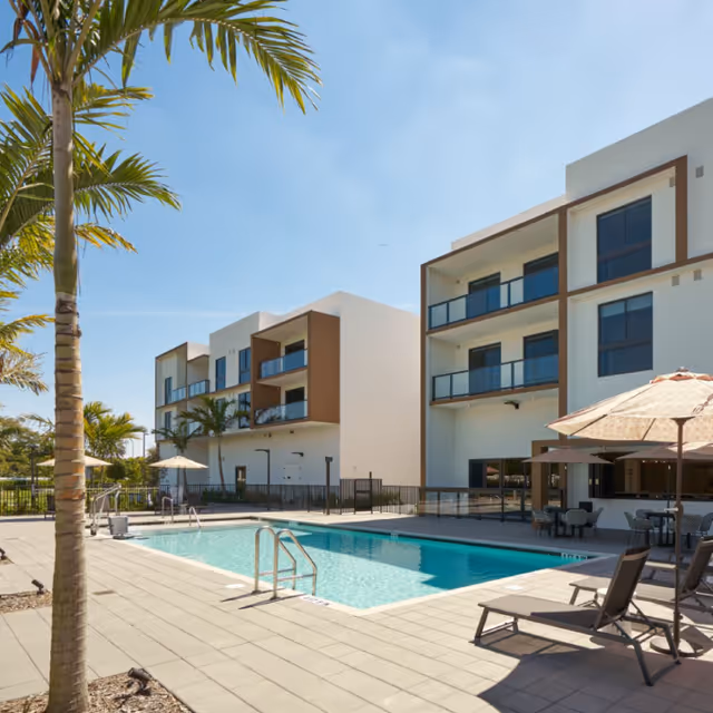 Outdoor swimming pool area at a senior living facility with lounge chairs, umbrellas, palm trees, and a modern three-story building in the background under a clear blue sky.
