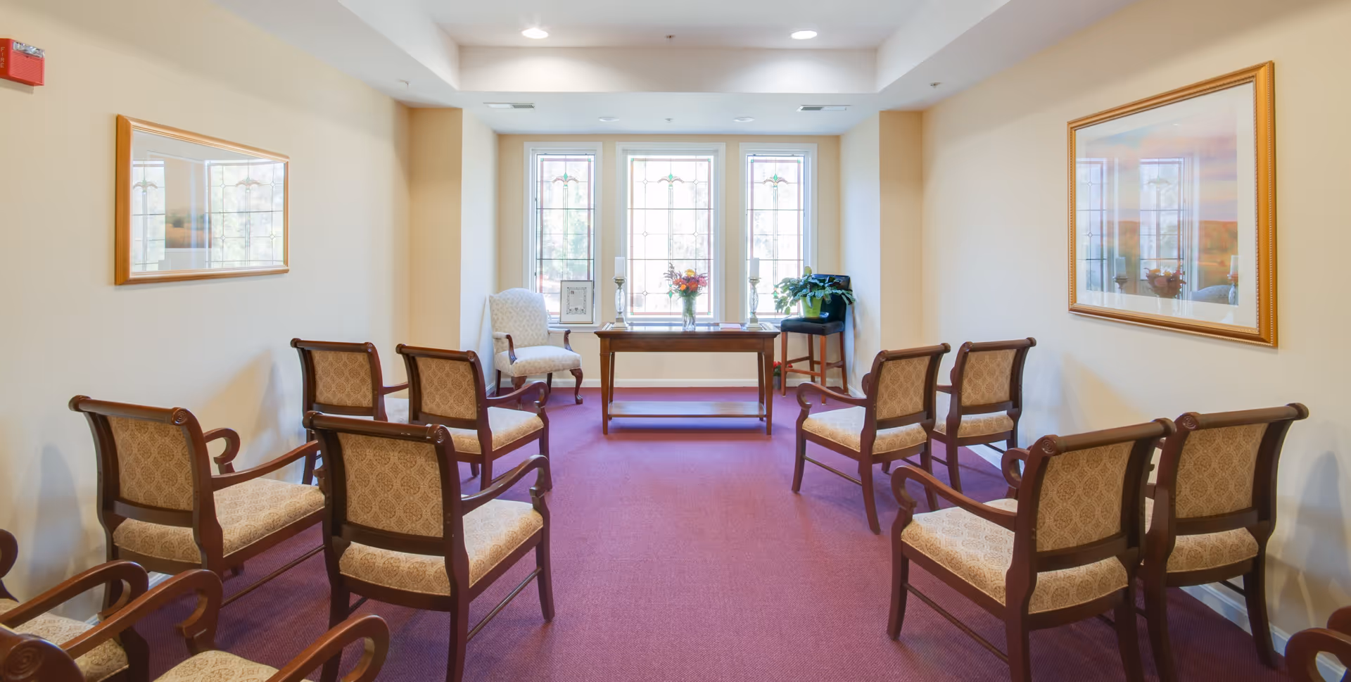 A bright interior meeting room at a senior living facility with upholstered chairs arranged facing a table in front of three tall windows.