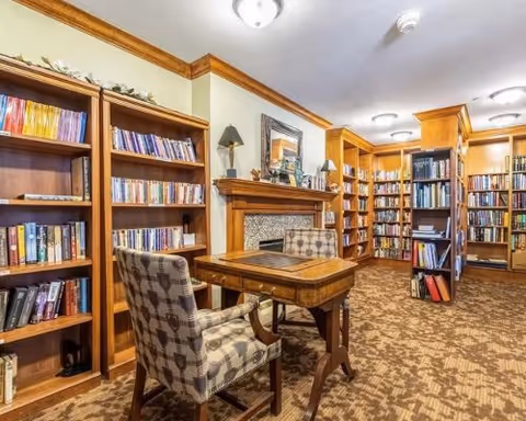 Cozy library room with wooden bookshelves filled with books, a wooden table with a patterned cushioned chair, a fireplace with a decorative mantle, and warm lighting from ceiling fixtures.