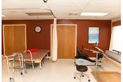 A physical therapy or rehabilitation room with a therapy table, walker, exercise equipment including a stationary bike, a stool, a clock on the wall, and a privacy curtain. The walls are painted brown and white, and there is a window with blinds allowing natural light into the room.