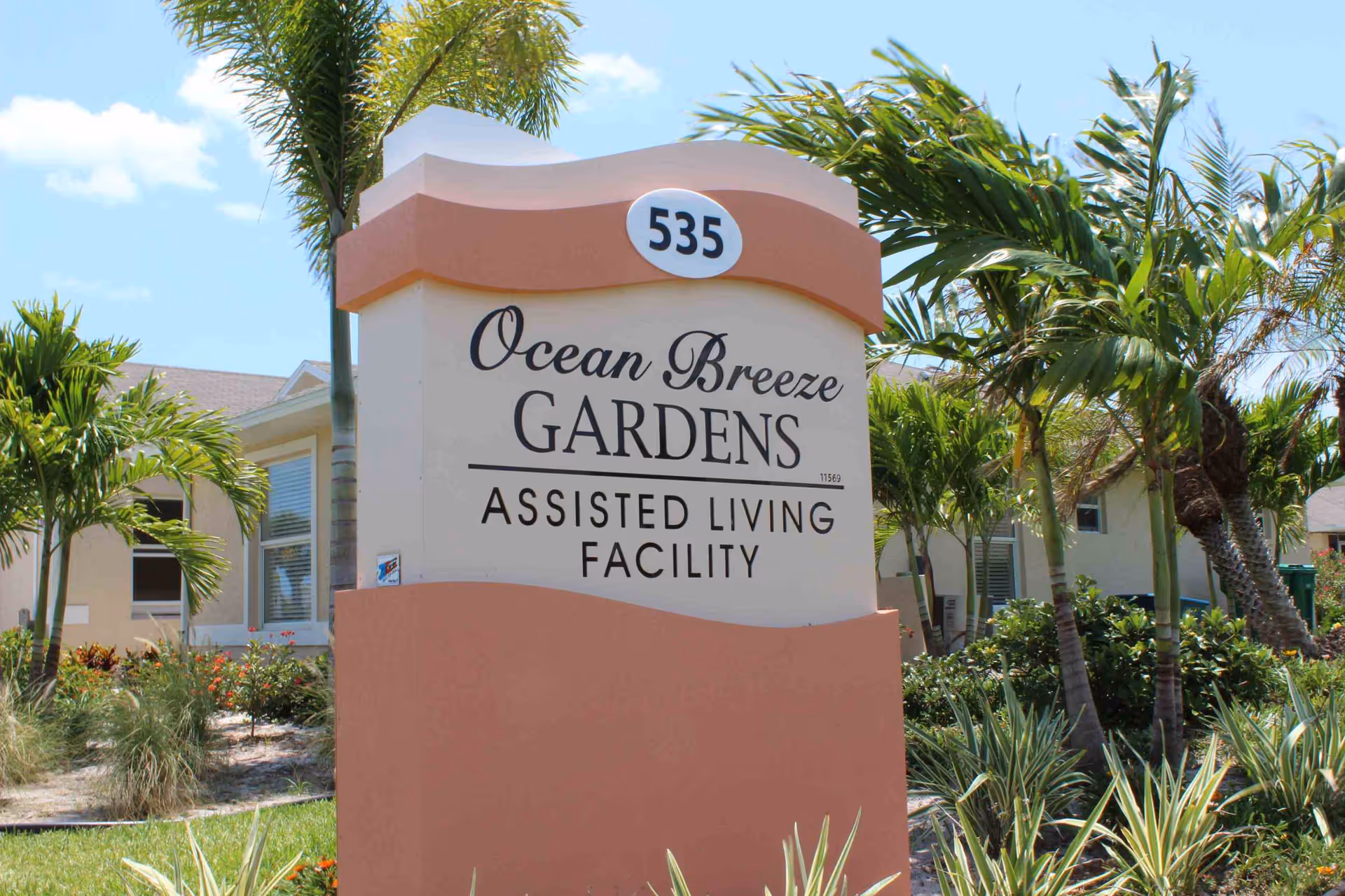 Outdoor view of the entrance sign for Ocean Breeze Gardens Assisted Living Facility surrounded by palm trees and landscaping under a clear blue sky.