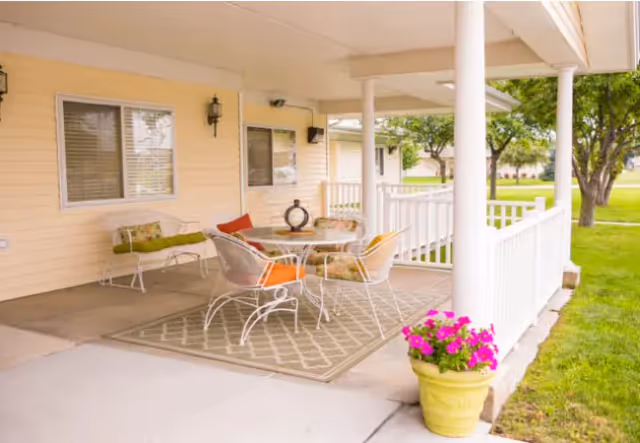 Covered outdoor patio area with white railing and columns, featuring a round glass table with four metal chairs with colorful cushions, a bench with cushions against the wall, a decorative lantern on the table, and a large yellow planter with pink flowers on the concrete floor. Green grass and trees are visible in the background.