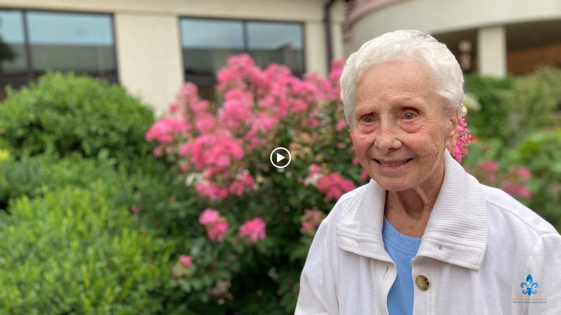 An elderly woman with white hair wearing a white jacket and blue shirt is smiling outdoors in front of green bushes and pink flowers, with a building visible in the background.