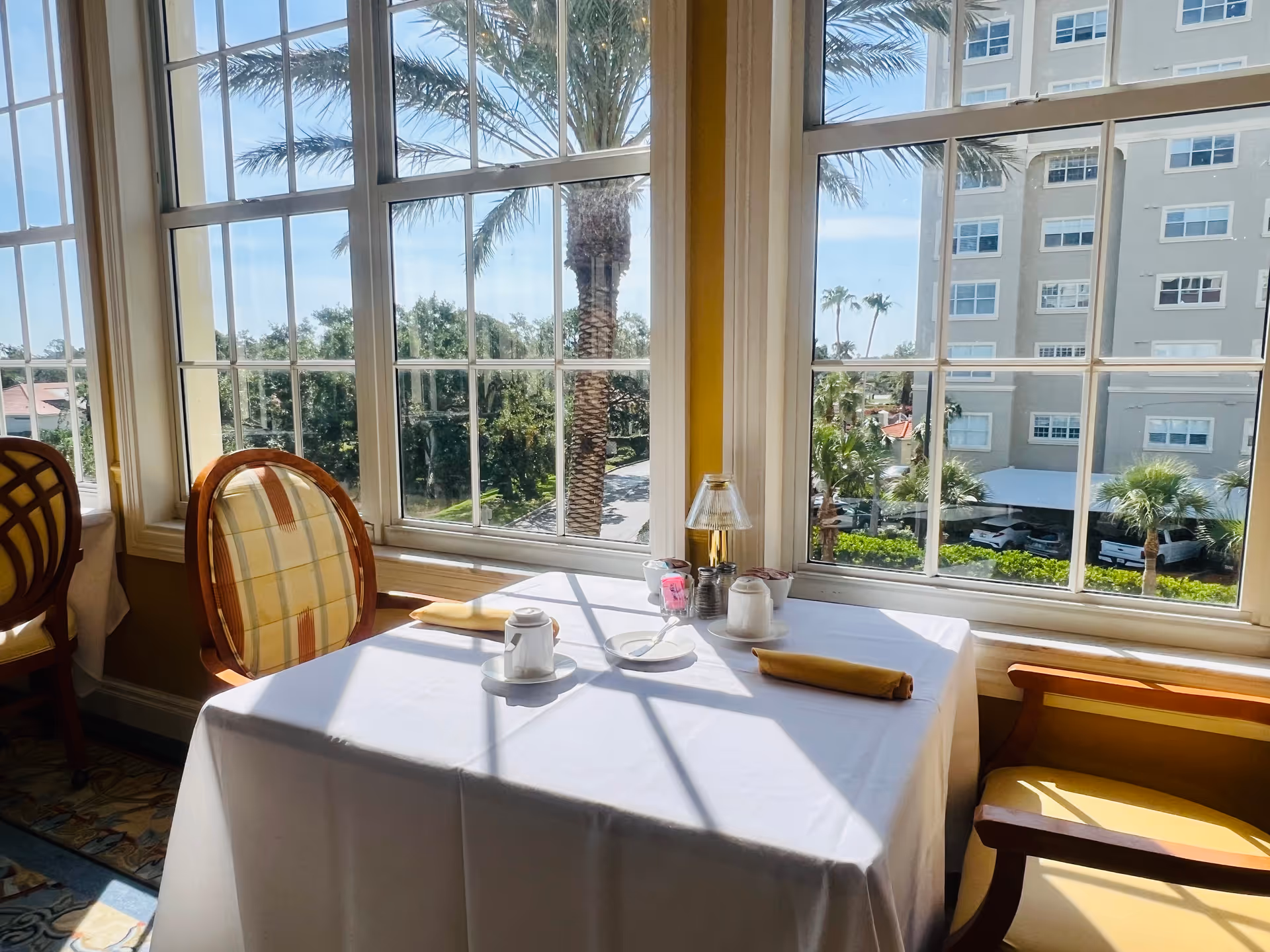 A dining table set for two by large windows with a view of palm trees and a multi-story building outside. The table is covered with a white tablecloth and has two yellow napkins, a small lamp, and condiments on it. Two chairs with patterned cushions are placed on either side of the table.