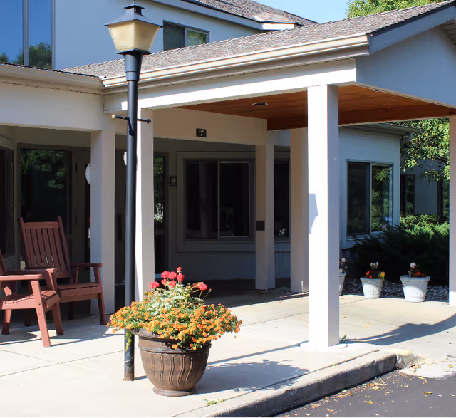 Covered entrance of a senior living facility with white columns, a lamppost, flower planters and wooden chairs.