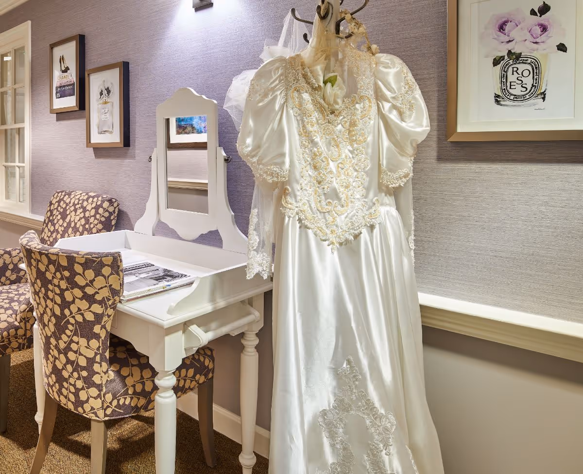 A vintage white satin dress with intricate lace detailing hangs on a hook next to a white vanity table with a mirror. Two patterned chairs are positioned near the table, and framed artwork decorates the lavender-colored wall behind them.