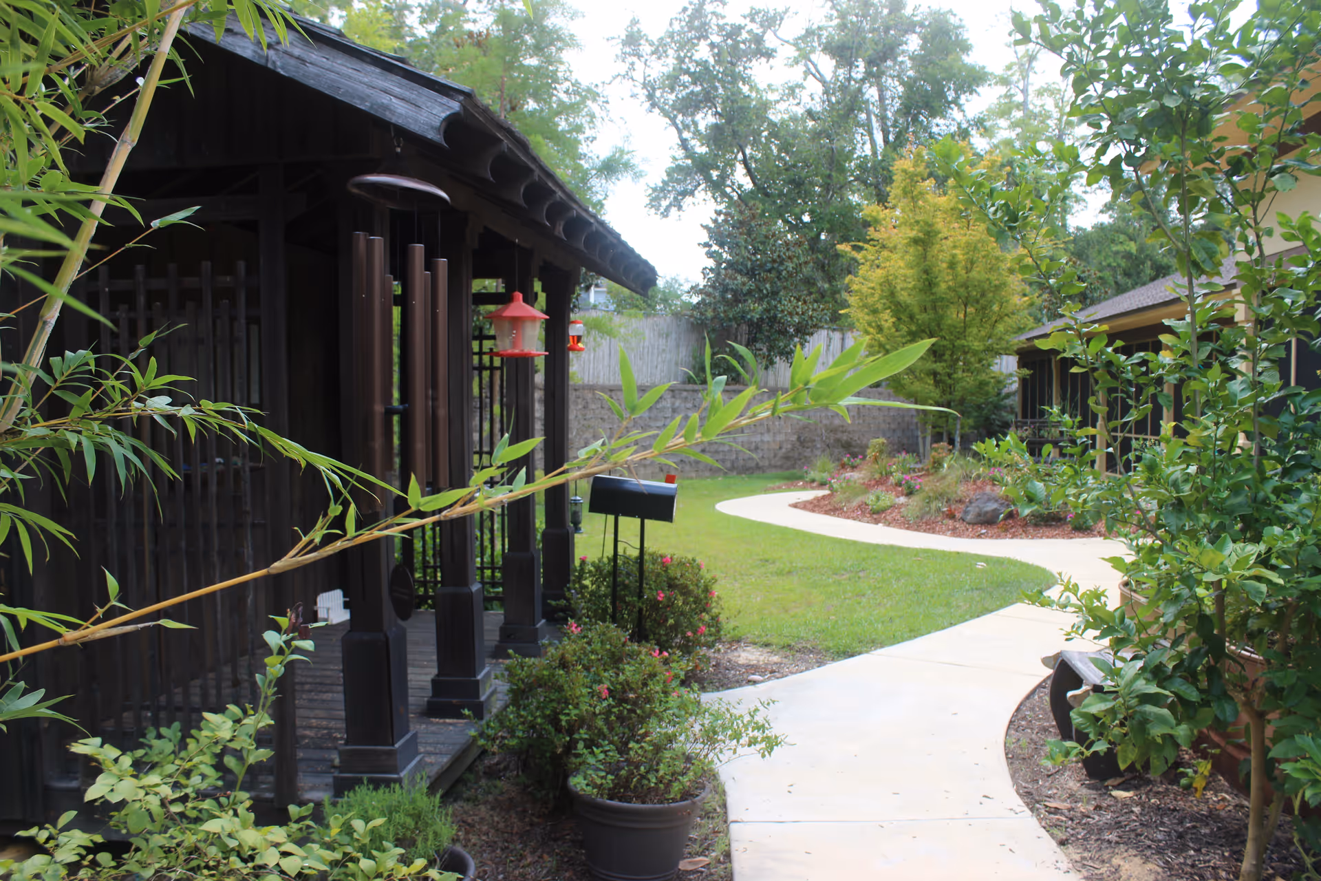 A garden area with a curved concrete pathway surrounded by green plants and trees. On the left side, there is a wooden structure with hanging wind chimes and bird feeders. The area appears peaceful and well-maintained.