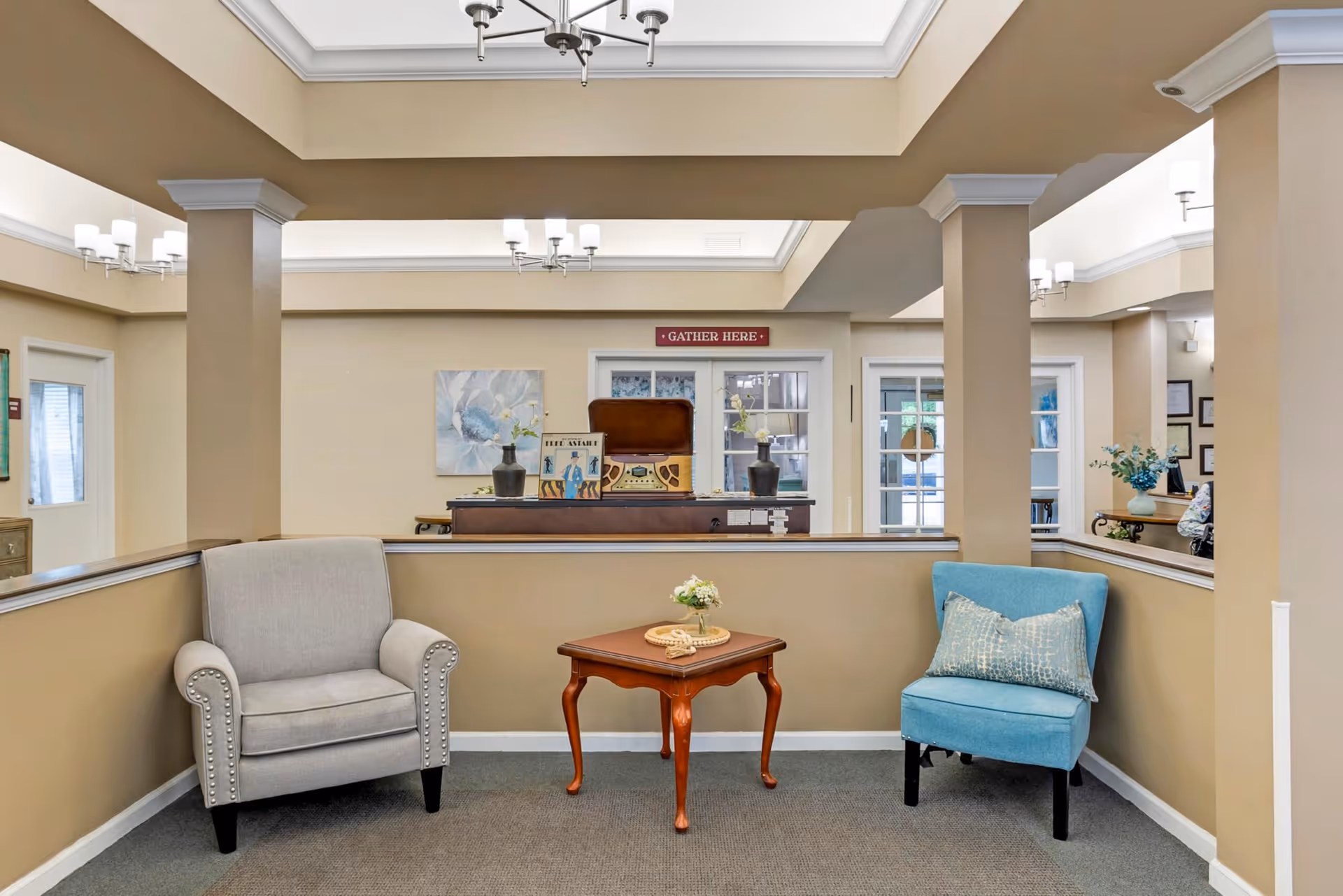 Lobby seating area with a gray armchair and a blue chair flanking a wooden side table under ceiling lights.