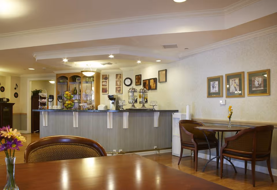 A cozy dining area in a senior living facility with wooden tables and chairs. The room features a counter with beverage dispensers, a coffee maker, and a fruit basket. The walls are decorated with framed pictures and a clock. There are flowers in vases on the tables, and the lighting is warm and inviting.