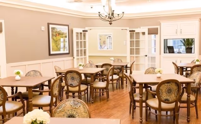 Bright dining room with multiple wooden tables and patterned chairs, small floral centerpieces, and a chandelier.