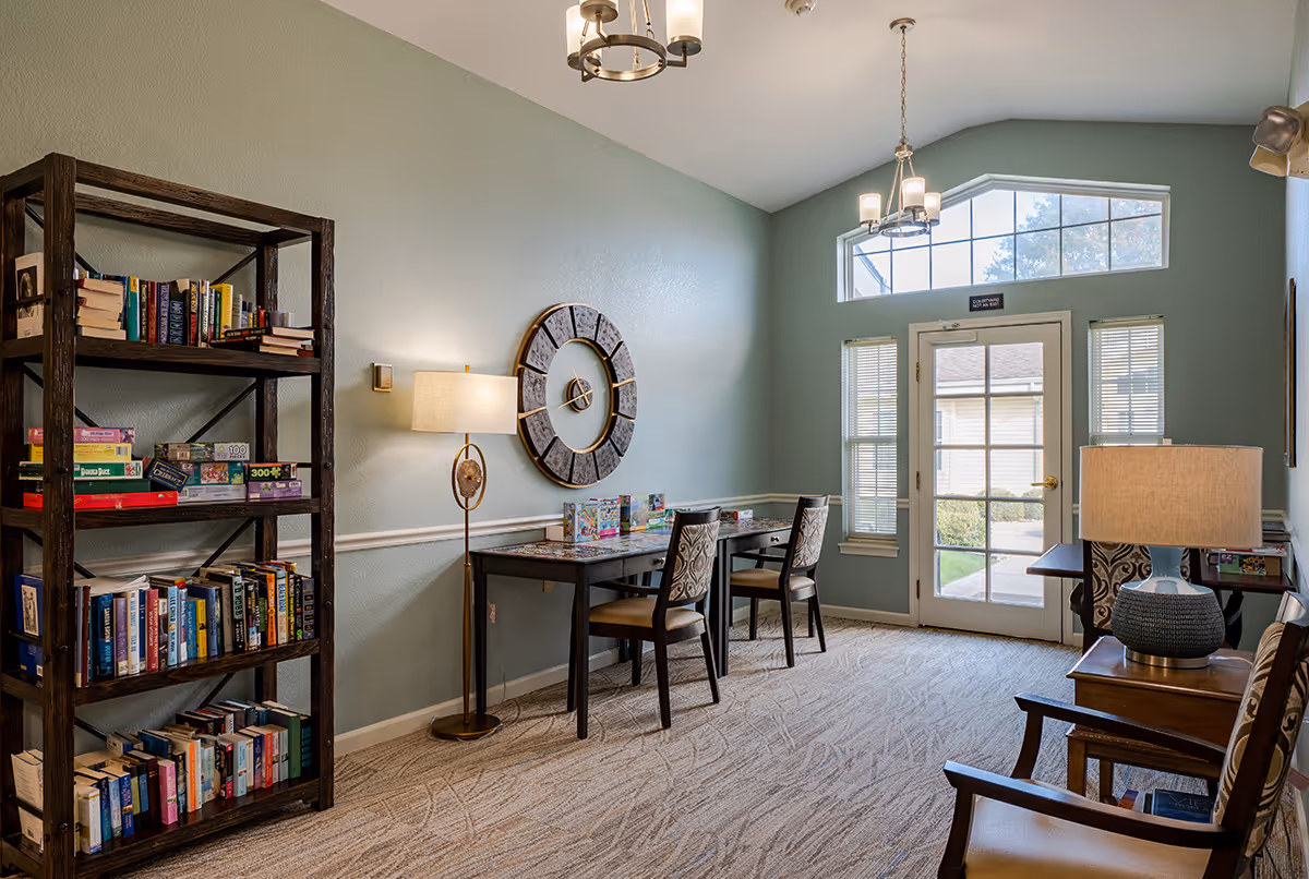 A cozy interior room with a bookshelf filled with books and board games on the left, two tables with chairs in the center, and a large decorative wall clock above the tables. There are two lamps, one floor lamp and one table lamp, providing warm lighting. The room has light blue walls, carpeted floor, and a door with windows leading outside, allowing natural light to enter.
